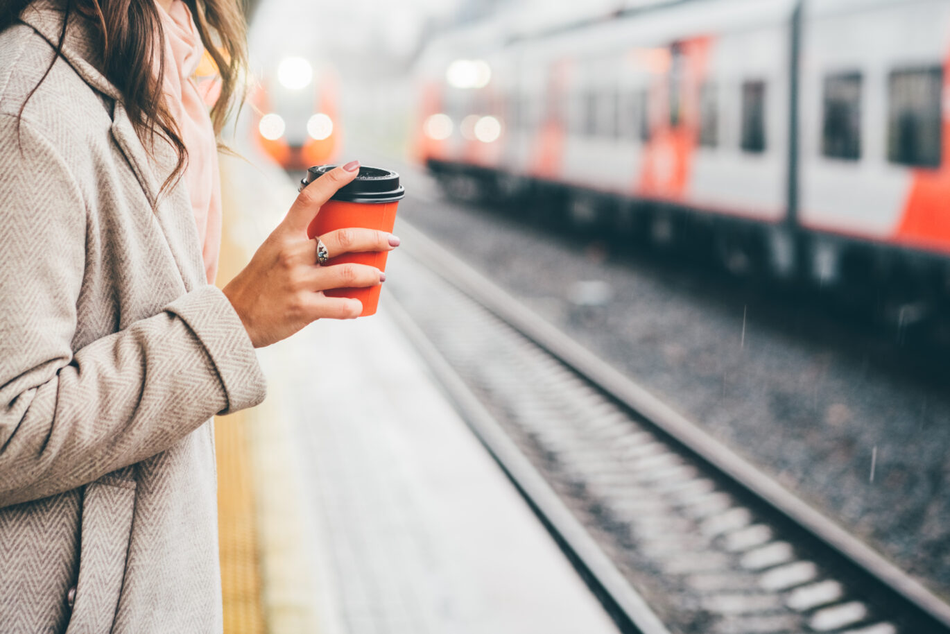 Een vrouw die een rode koffie to go beker vasthoudt terwijl ze staat te wachten op de trein